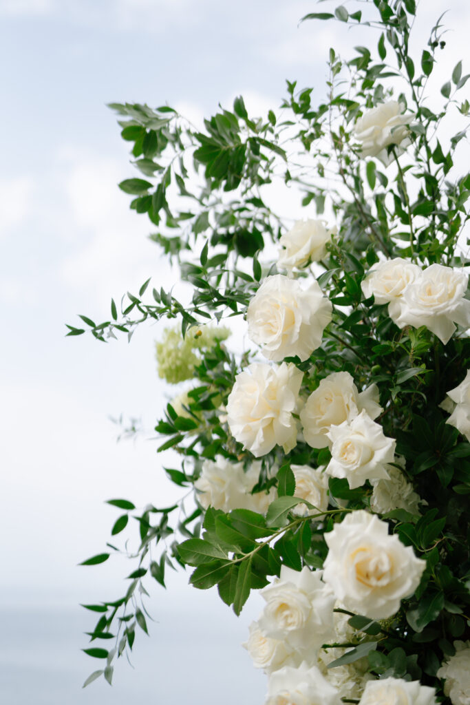 A lush arrangement of white roses and green foliage set against a bright, cloudy sky, creating a fresh and elegant outdoor atmosphere.