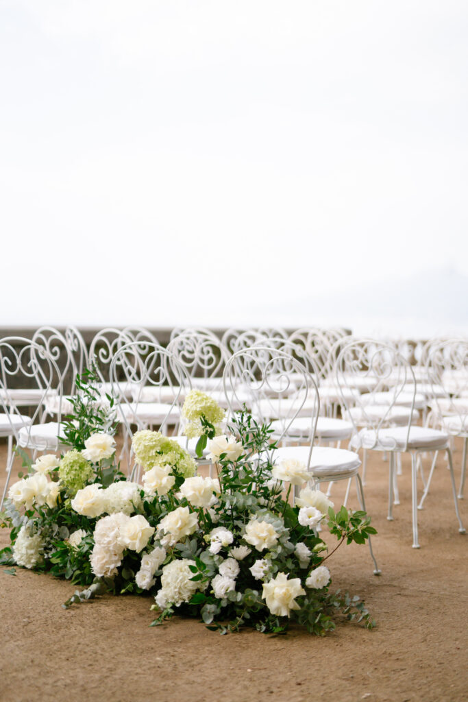 Rows of white ornate chairs are set up outdoors on sandy ground, with lush white and green floral arrangements placed in front, creating an elegant and airy ceremony setting.