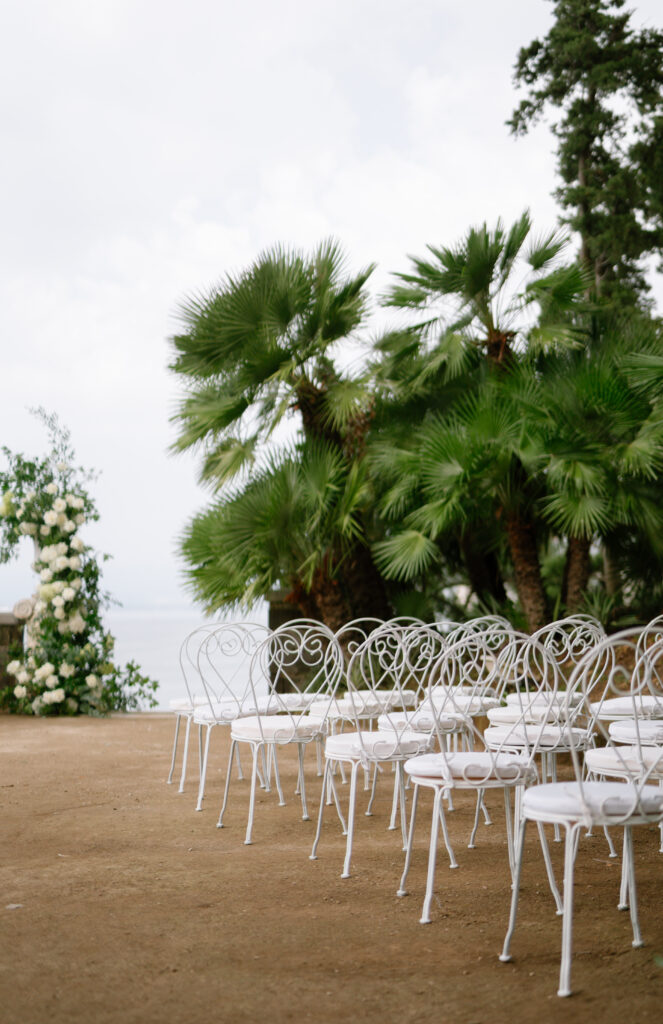 Rows of white metal chairs are arranged outdoors on sandy ground, facing a floral arch with white flowers and greenery. Tall palm trees and dense plants create a lush backdrop, suggesting a garden or beachside event.