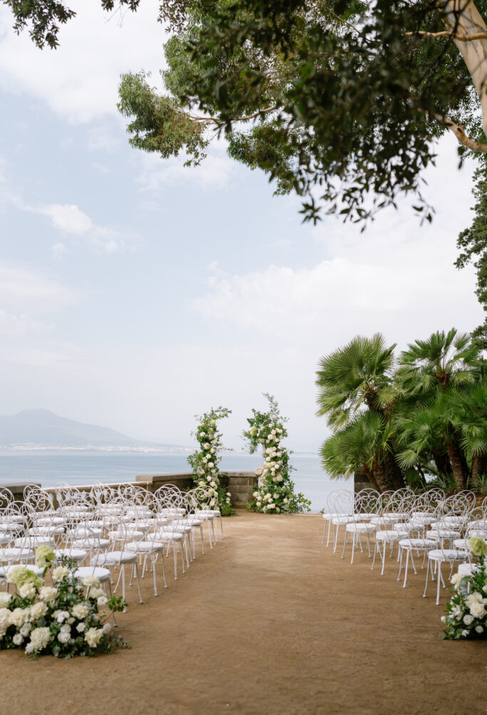 A wedding ceremony setup outdoors with white chairs arranged in rows facing a floral altar, surrounded by greenery, and overlooking the sea under a partly cloudy sky.
