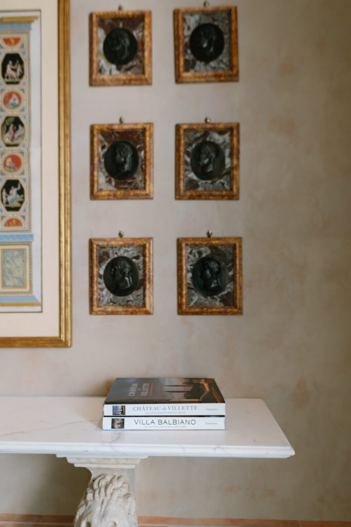 A marble table with two stacked books stands against a beige wall adorned with six framed round medallion artworks arranged in two columns. Part of a large, colorful framed print is visible on the left.