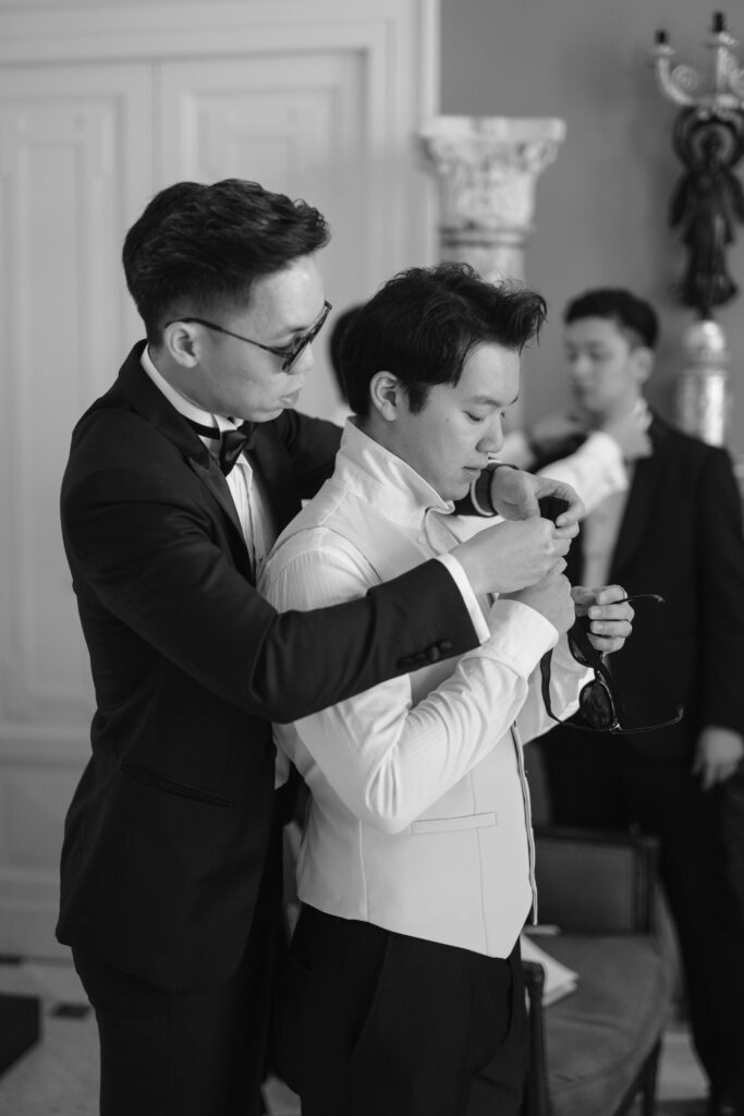 Two men in formal wear are getting ready; one helps the other adjust his bow tie, while a third man in the background also adjusts his tie. The photo is in black and white.