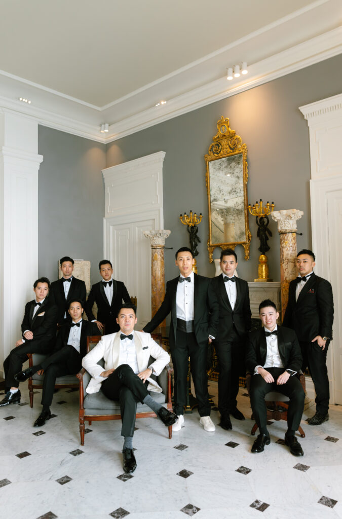 A group of eight men dressed in black tuxedos pose in an elegant room with white walls, marble columns, a gold-framed mirror, and classical decor. Four men are seated while the others stand behind them, all looking at the camera.