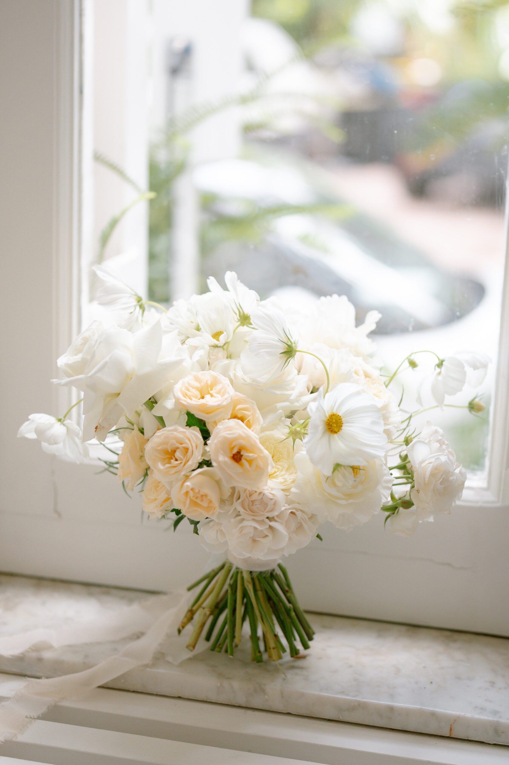 A bouquet of white and pale peach flowers with green stems rests on a marble windowsill in soft natural light, with a blurred outdoor scene visible through the window behind.