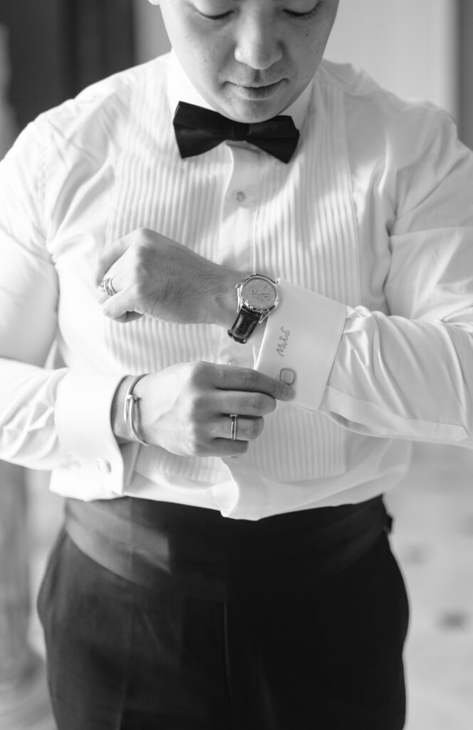 A man in formal attire adjusts his cuff while wearing a bow tie and a wristwatch. The photo is in black and white, focusing on his torso and hands as he prepares for a formal event.