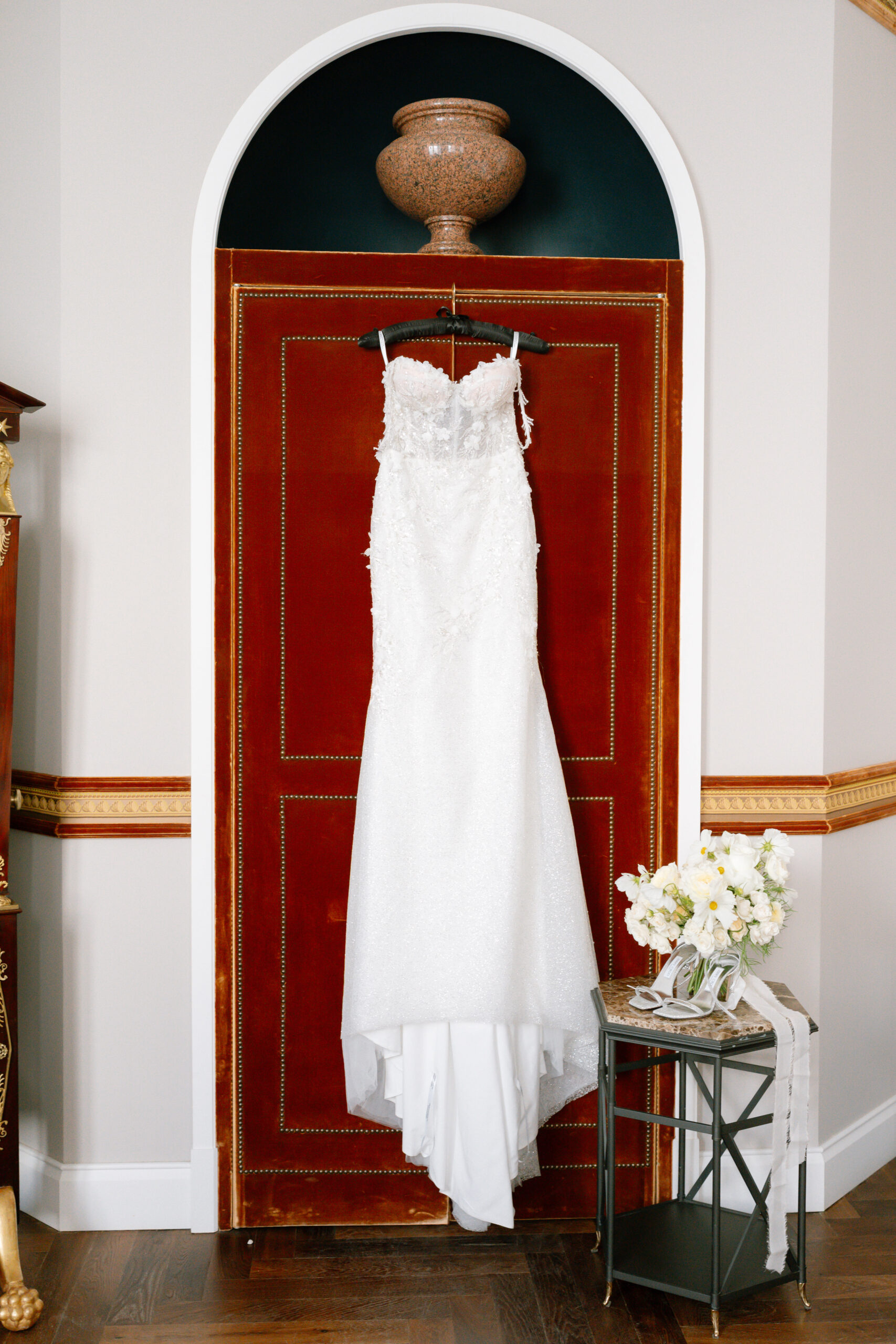 A white lace wedding dress hangs on a dark red velvet door beneath a decorative urn. A small table with a bouquet of white and yellow flowers sits to the right.
