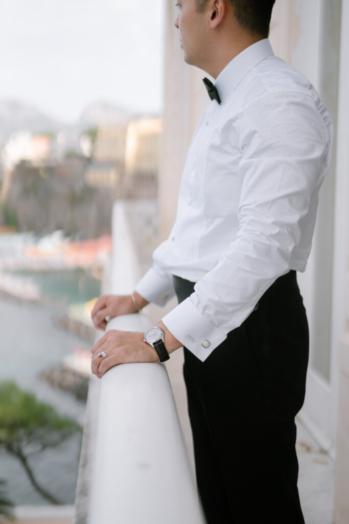 A man in a white dress shirt, black bow tie, and black pants stands on a balcony overlooking a scenic, coastal view. He wears a watch and wedding ring, gazing thoughtfully into the distance.