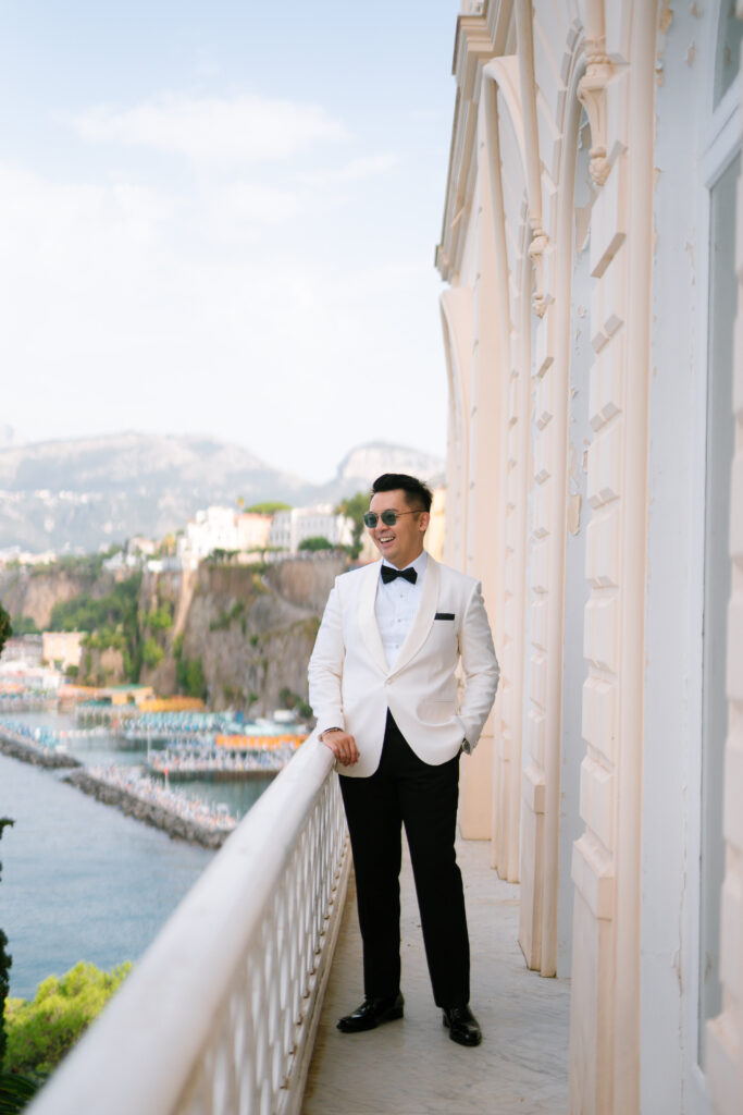 A man in a white tuxedo jacket, black bow tie, and sunglasses stands smiling on a balcony overlooking a coastal town with cliffs, buildings, and the sea in the background.