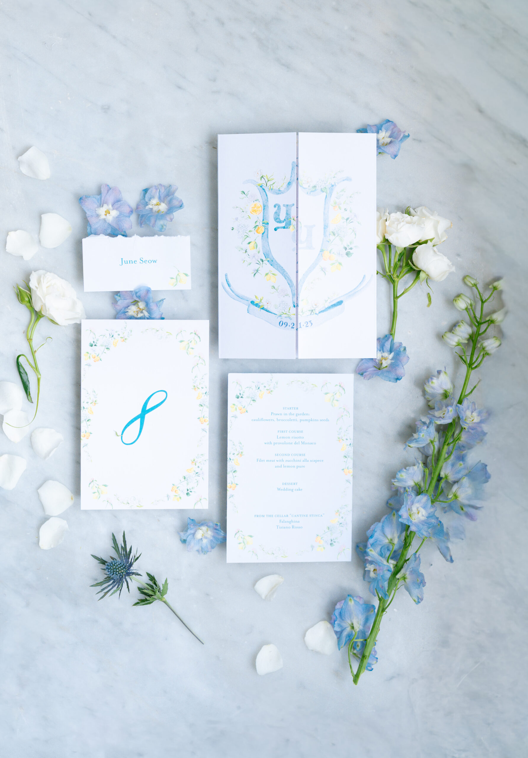 Flat lay of elegant wedding stationery featuring invitations, a name card, and a program surrounded by white petals, blue delphiniums, and white lisianthus flowers on a light marble surface.