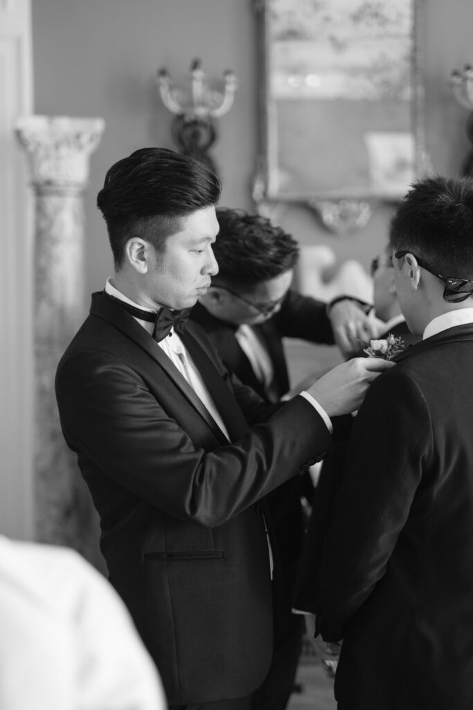Three men in tuxedos adjust each others boutonnieres in a well-decorated room. The image is black and white, and the atmosphere appears formal, likely in preparation for a special event such as a wedding.