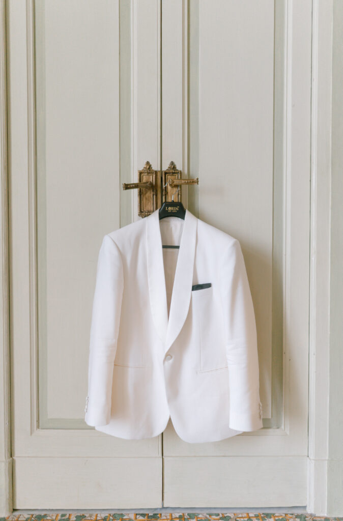 A white suit jacket with a black pocket square hangs on a black hanger from a brass handle, against a pair of cream-colored paneled doors.