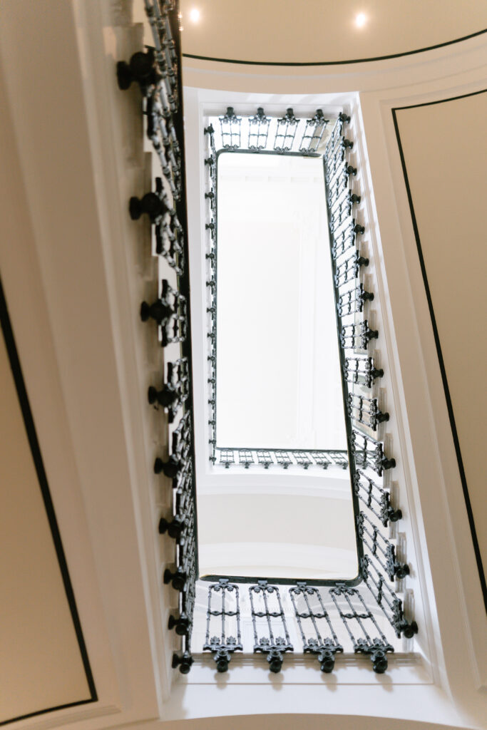 A view looking up through the rectangular center of a spiral staircase with ornate black railings and white walls, showing multiple levels leading upwards.