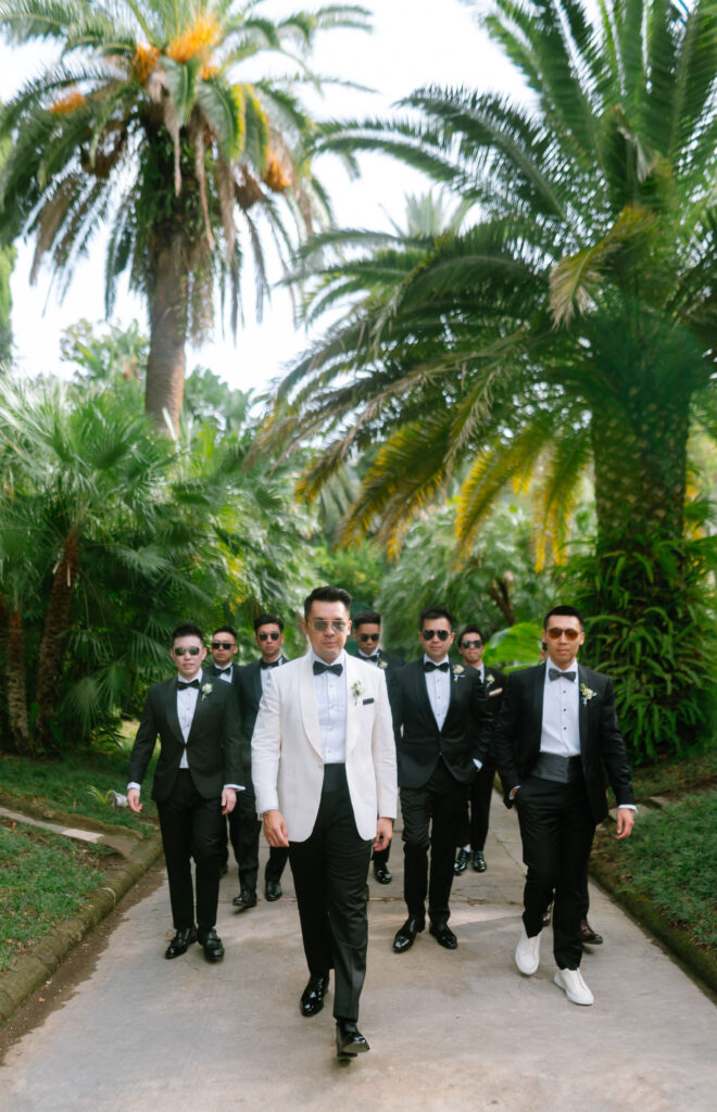 A group of men in tuxedos, one wearing a white jacket and the others in black, walk confidently down a palm-lined path outdoors. All wear sunglasses and formal shoes, creating a stylish and coordinated appearance.