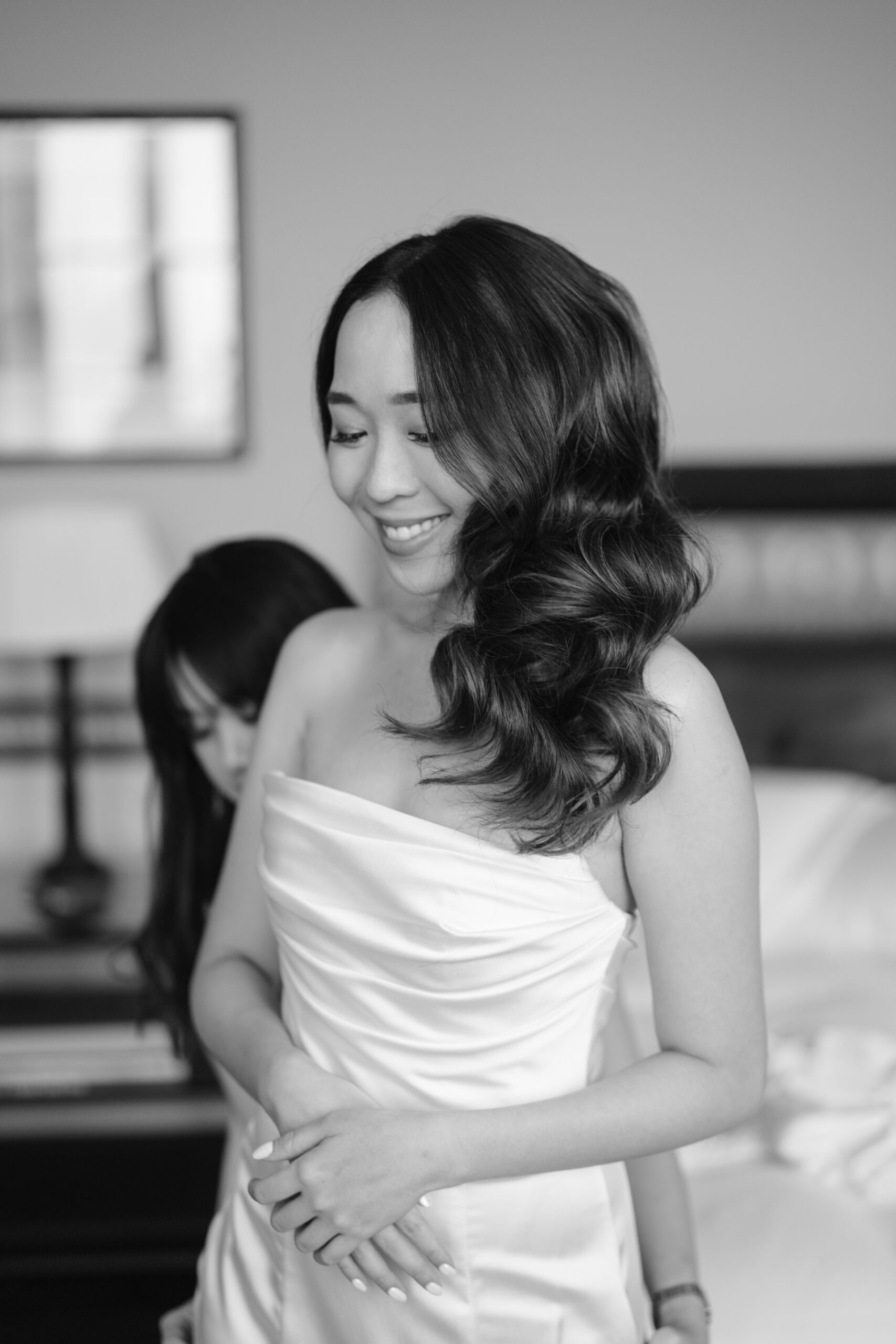 A smiling woman in a strapless dress stands indoors as someone helps her from behind. The scene is in black and white, with soft lighting and a blurred background.