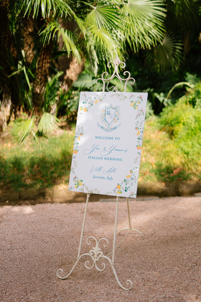 A decorative wedding welcome sign on an ornate white stand, set outdoors among lush green plants. The sign reads, Welcome to John & Gemmas Italian Wedding, Villa Astor, Sorrento, Italy, with floral designs around the border.
