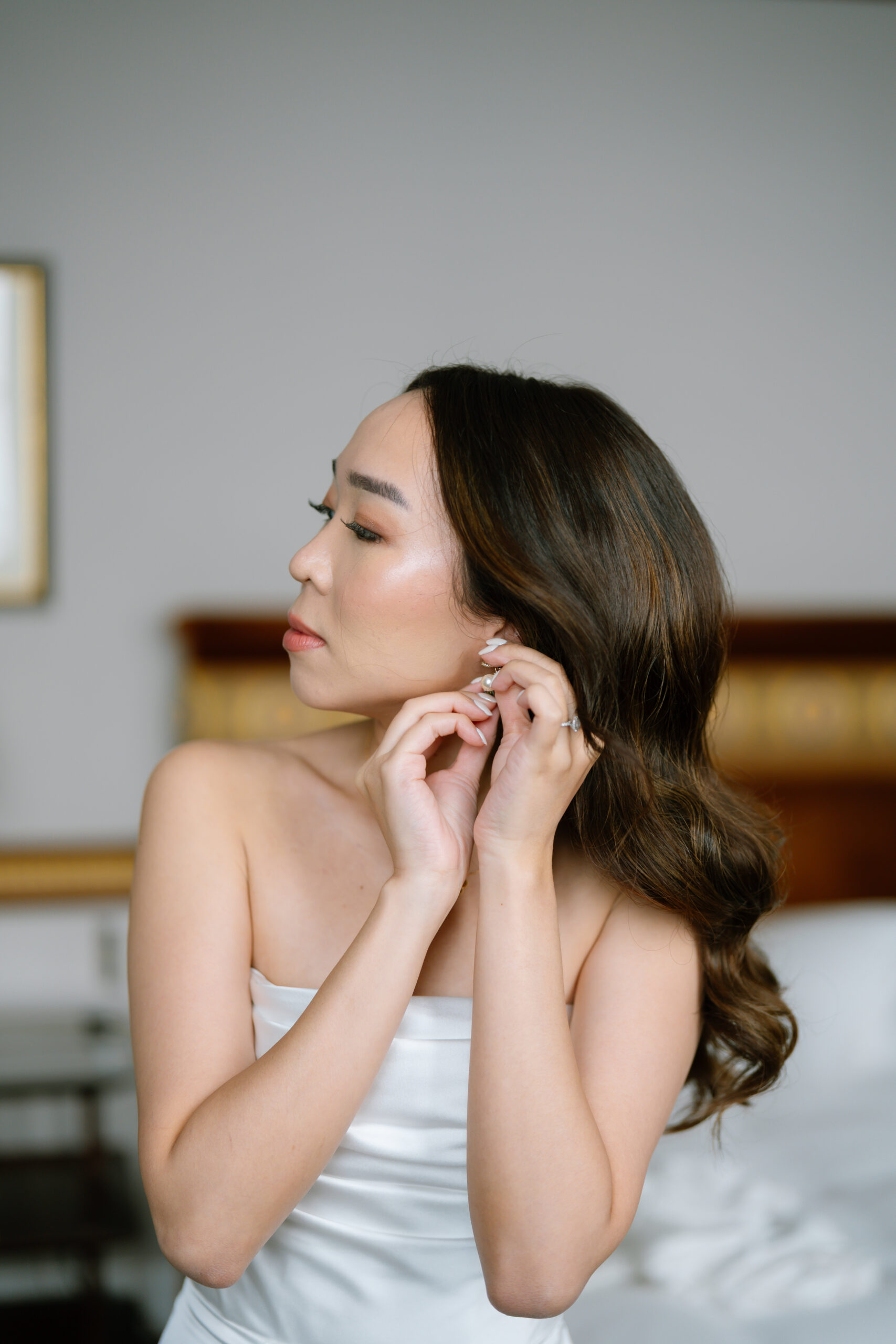A woman with long wavy hair, wearing a white strapless top, is putting on an earring. She is looking to the side and standing indoors in a softly lit room with a blurred background.