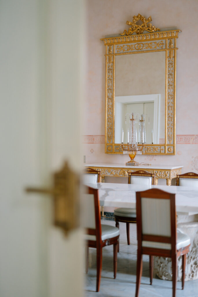 A view into an elegant room with a marble table, wooden chairs with white cushions, and a large ornate gold mirror above a matching console table. The foreground shows a partially open door with a brass handle.