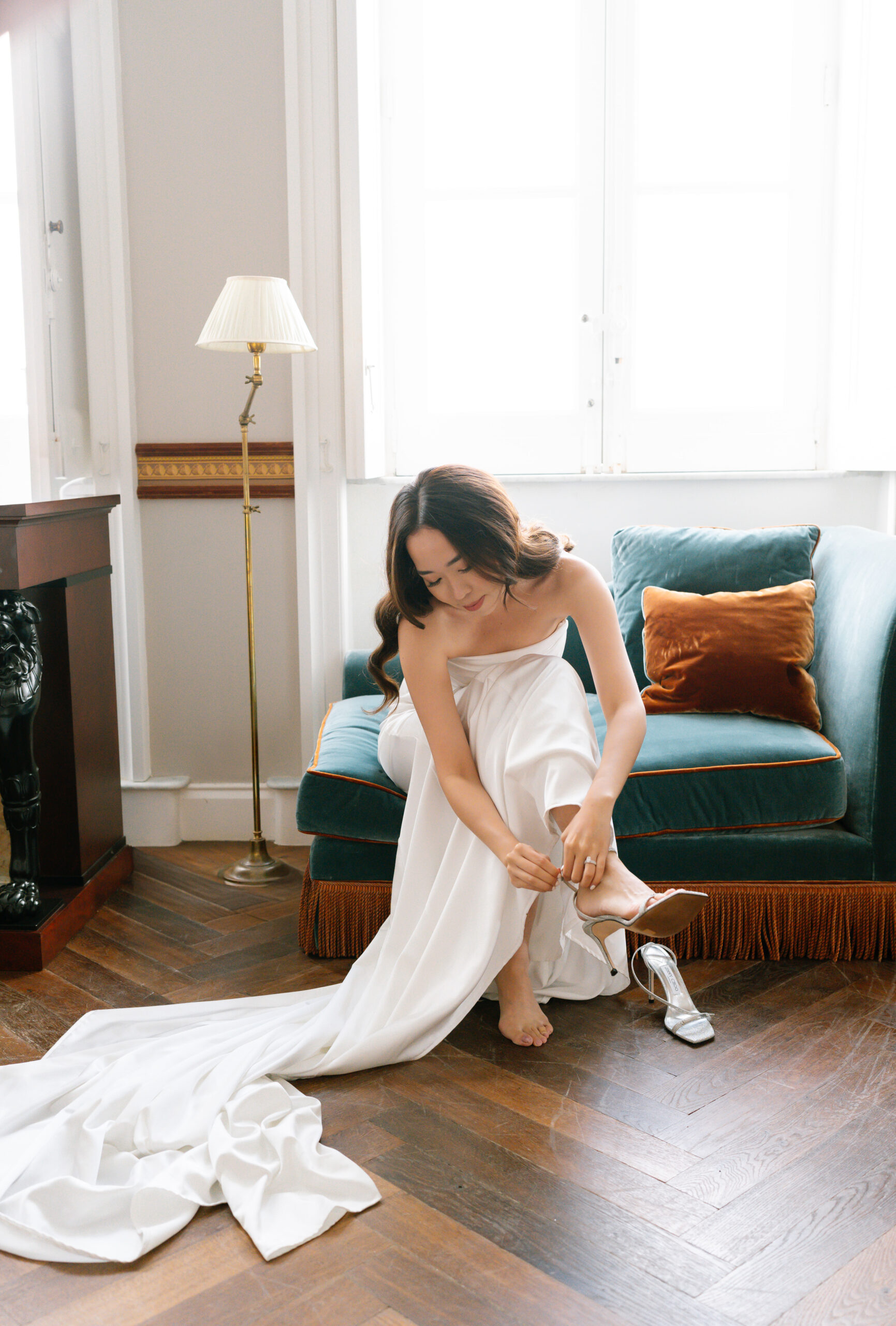 A woman in a strapless white gown sits on a green velvet sofa, adjusting her shoe. One silver shoe is off to the side. Soft natural light streams in from tall windows behind her.