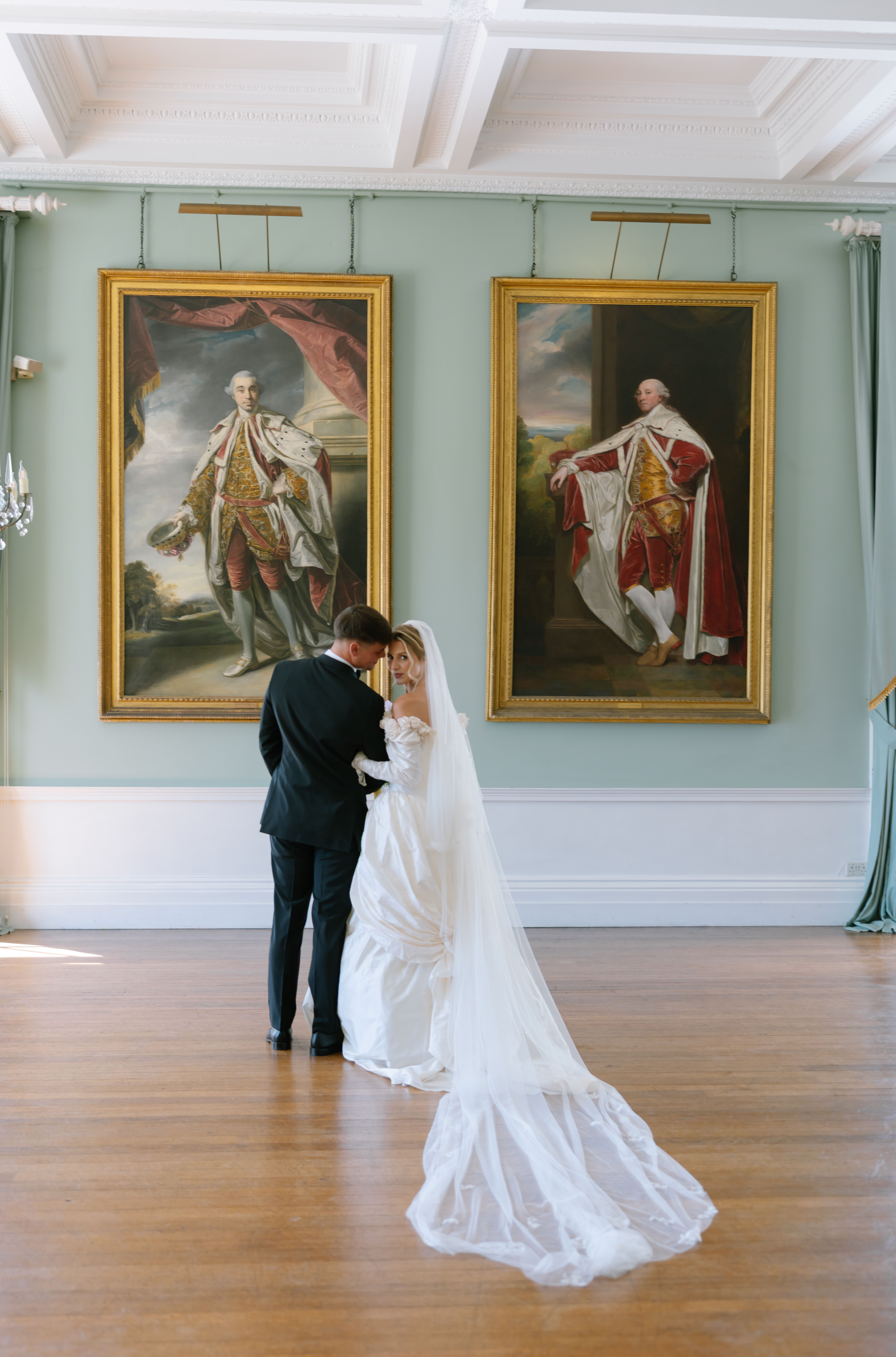 A bride and groom stand together in a grand room with wooden floors, large windows, and two historical portraits in ornate frames hanging on a pastel green wall.