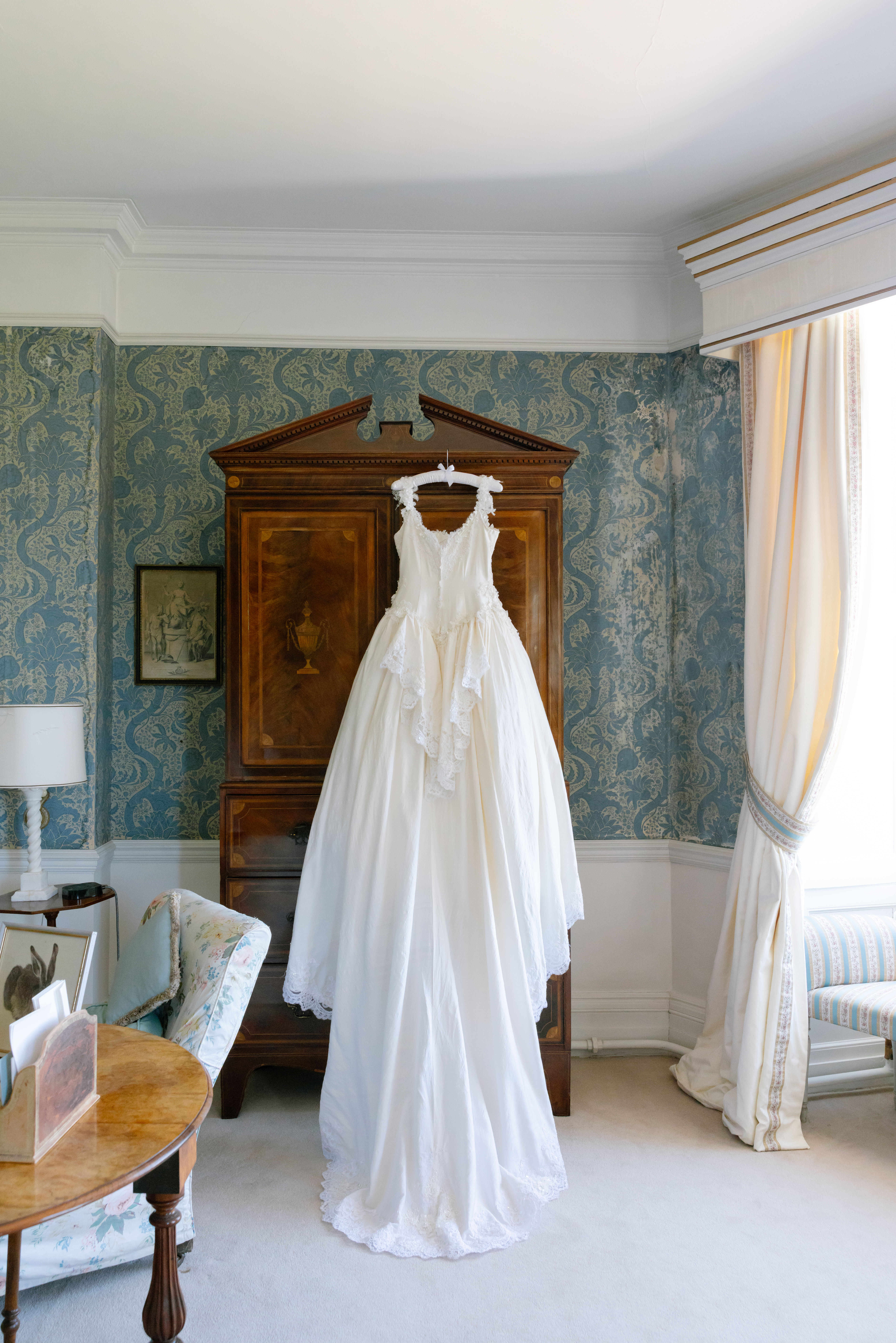 A white wedding dress hangs on an ornate wooden wardrobe in a vintage-style room with patterned blue wallpaper, a floral chair, a wooden table, and a window with flowing curtains.
