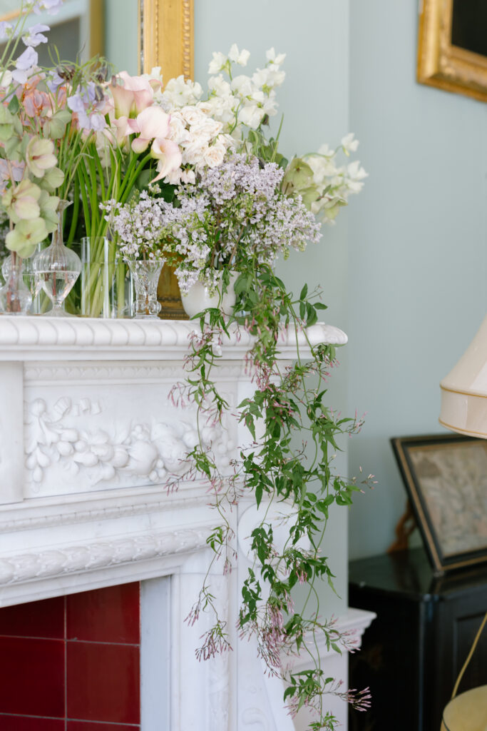 A white marble mantel with various vases of pastel flowers and trailing greenery, set against a pale wall with a gold-framed mirror above.