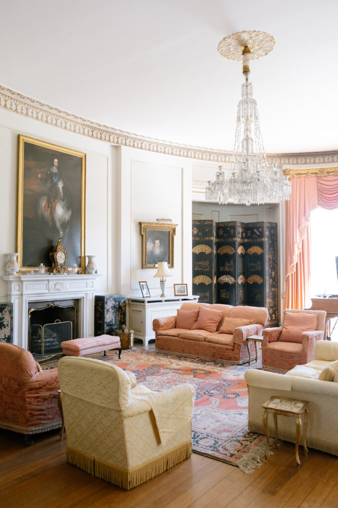 Elegant living room with ornate chandelier, vintage sofas, fireplace, framed portraits, decorative screen, large rug, and tall windows covered by pink drapes. Classic, historical decor with gold and white accents throughout.