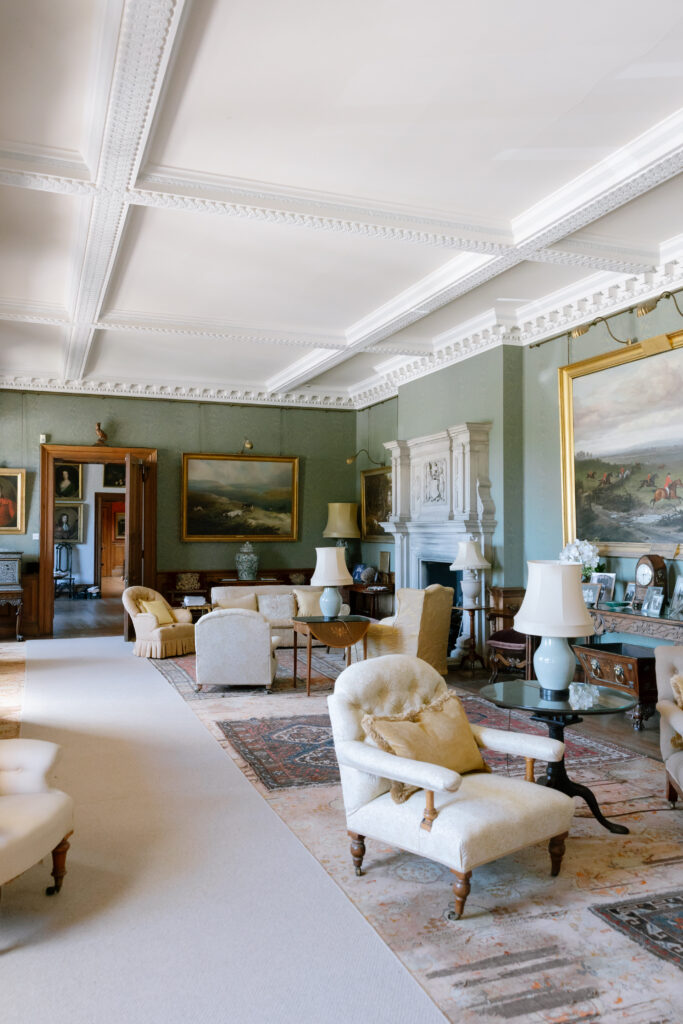 Elegant, spacious living room with vintage armchairs, ornate fireplace, large paintings, and lamps on tables, set on patterned rugs and carpet, with green walls and decorative ceiling moldings.
