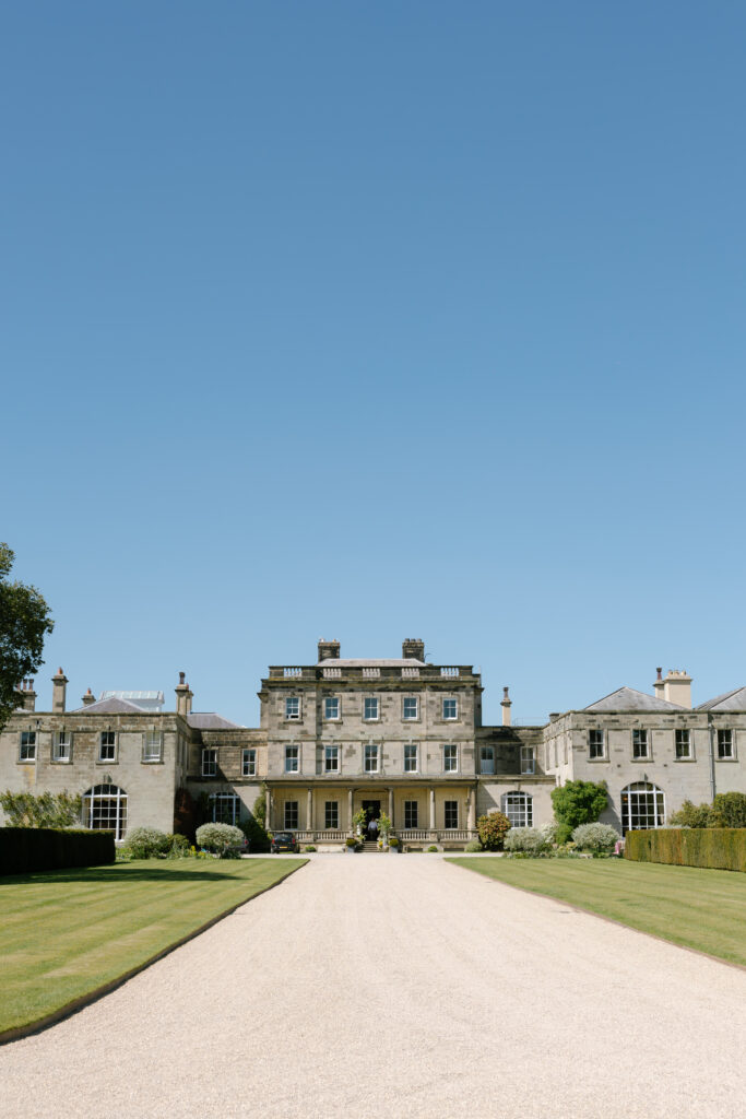 A grand stone mansion with symmetrical wings, large windows, and manicured lawns, sits under a clear blue sky at the end of a long gravel driveway.