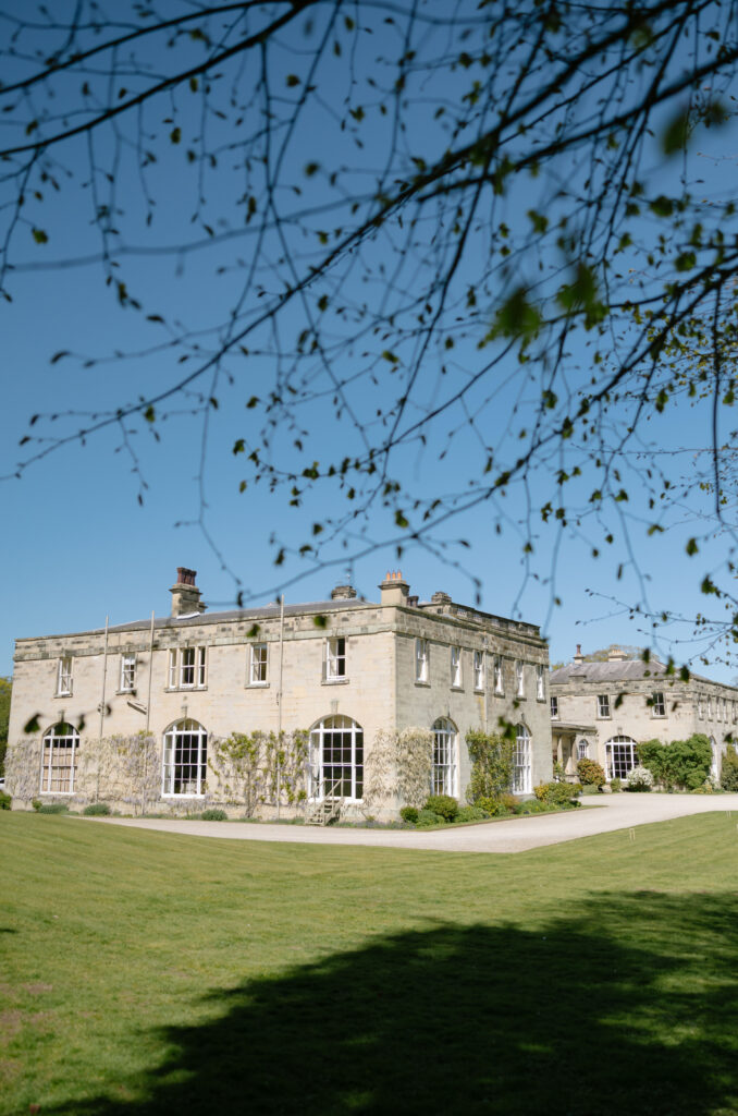 A large stone mansion with tall windows stands under a clear blue sky, framed by delicate tree branches and surrounded by a well-kept green lawn.