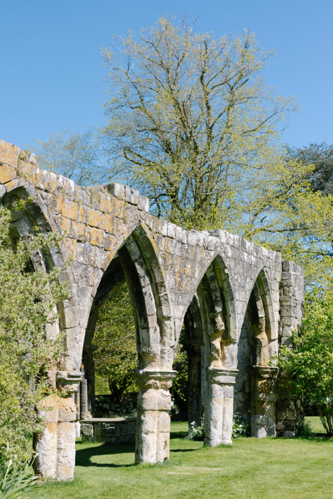 Stone arches of ancient ruins stand on green grass, surrounded by trees with fresh spring leaves under a clear blue sky.