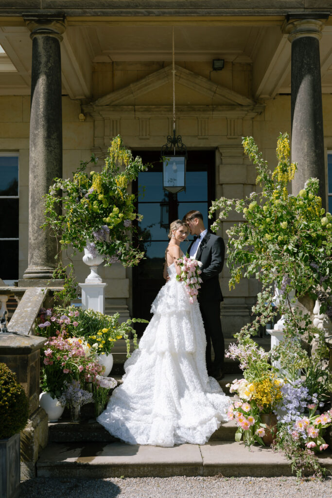 A bride and groom stand on the steps of a grand building, surrounded by lush floral arrangements, sharing an intimate moment on their wedding day.