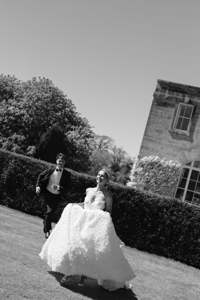 A bride in a white dress runs joyfully on grass, followed by a groom in a suit. They are outdoors near a large hedge and an old stone building under a clear sky.