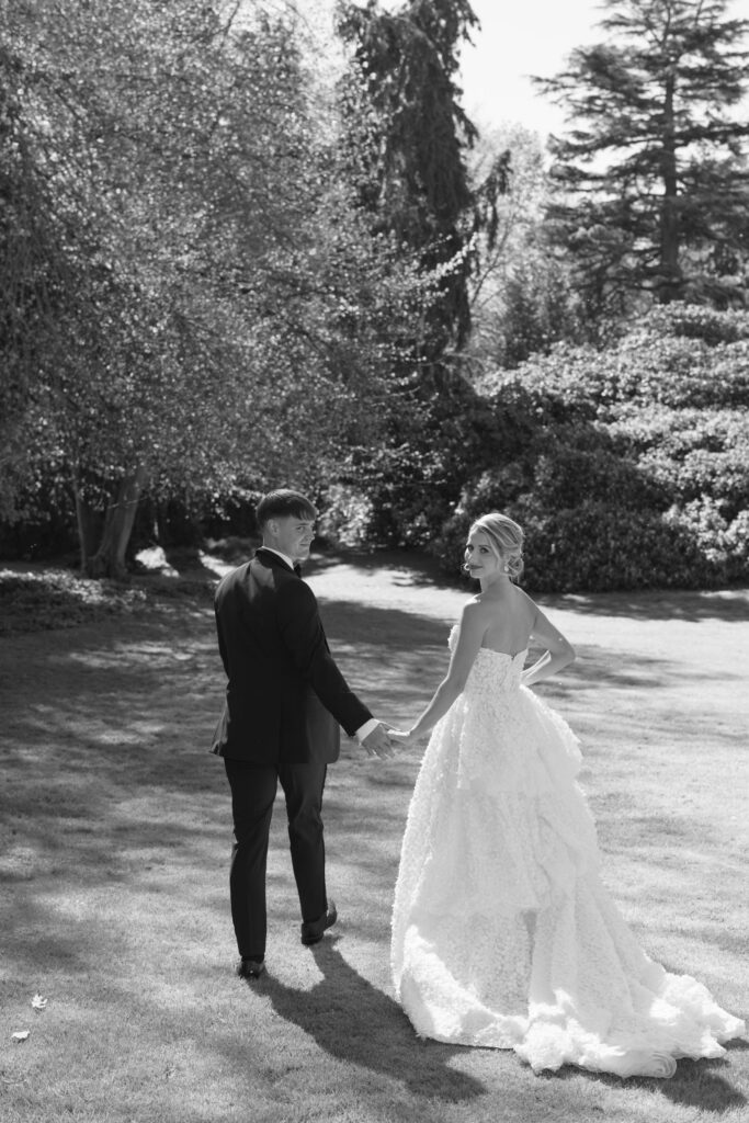 A bride and groom walk hand in hand on a grassy lawn. The bride looks back and smiles, wearing a strapless gown, while the groom wears a suit. Trees and sunlight fill the background.