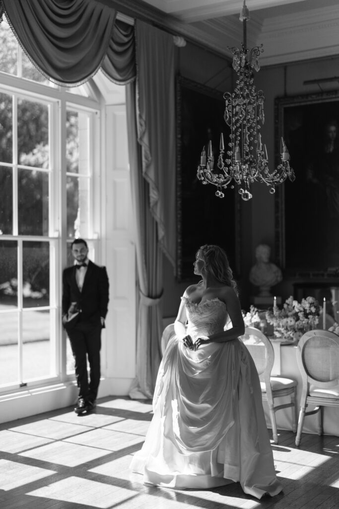 A bride in a wedding gown stands near a dining table in a sunlit, elegant room; a groom in a suit stands by a large window in the background. The scene is in black and white.