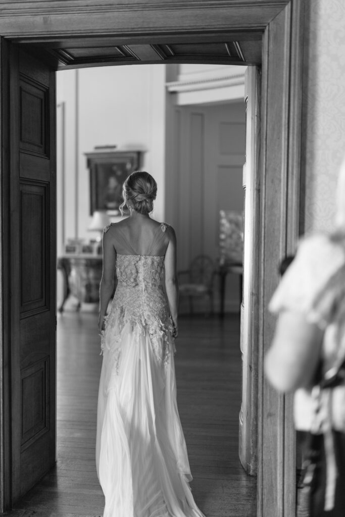 A bride in a lace wedding dress stands in a doorway, facing away, entering a bright, elegant room. The image is black and white.