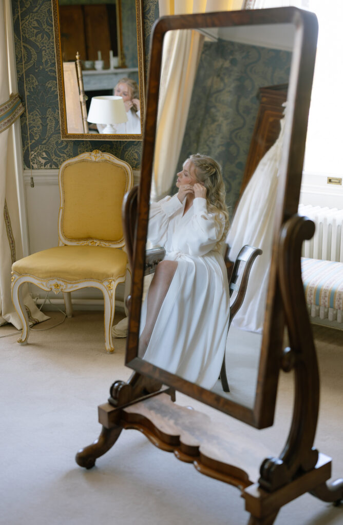A woman in a white dress sits on a chair, seen through a tall mirror in an elegant, vintage-style room with ornate furniture and patterned wallpaper.