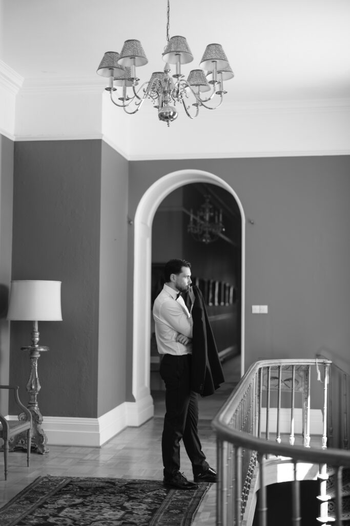 A man in formal attire stands by a railing in a stylish hallway, holding a jacket over his shoulder. The scene is in black and white, with elegant decor, a chandelier, and an arched doorway in the background.