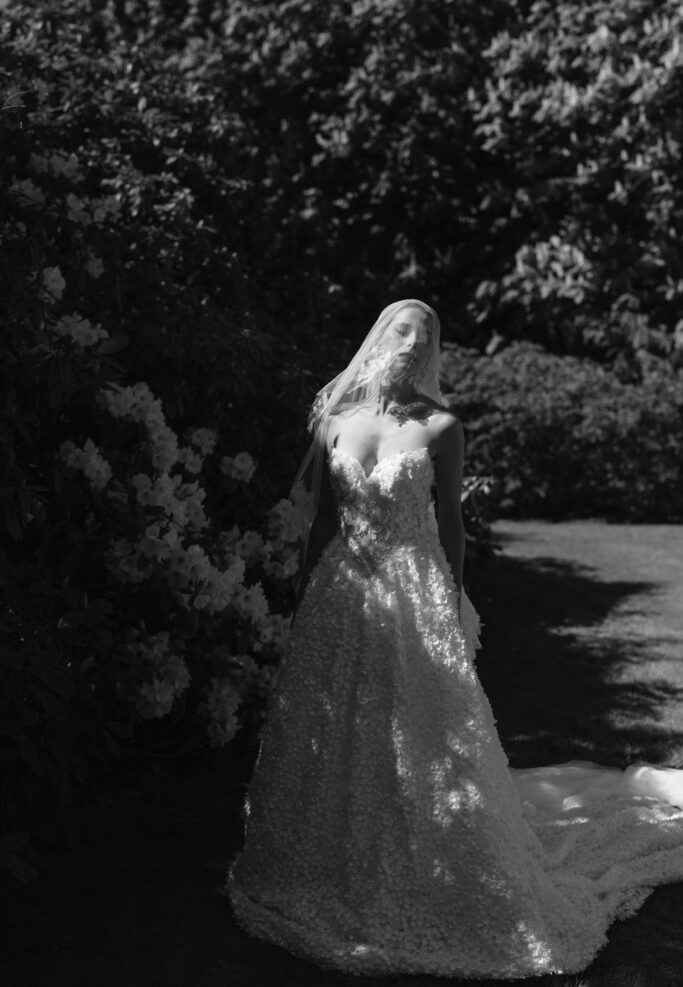 A bride in a strapless, textured gown and veil stands outdoors in sunlight, surrounded by lush bushes and flowers, with dramatic shadows cast across her dress and face.