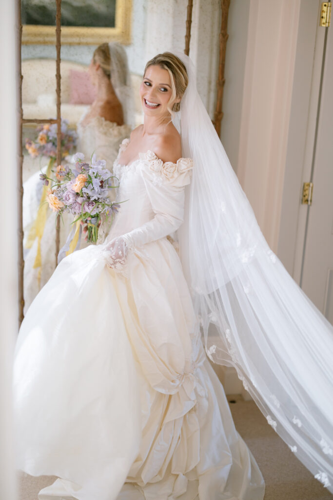 A smiling bride in an off-the-shoulder white wedding dress and long veil holds a bouquet of pastel flowers, standing indoors near a mirror reflecting her image.