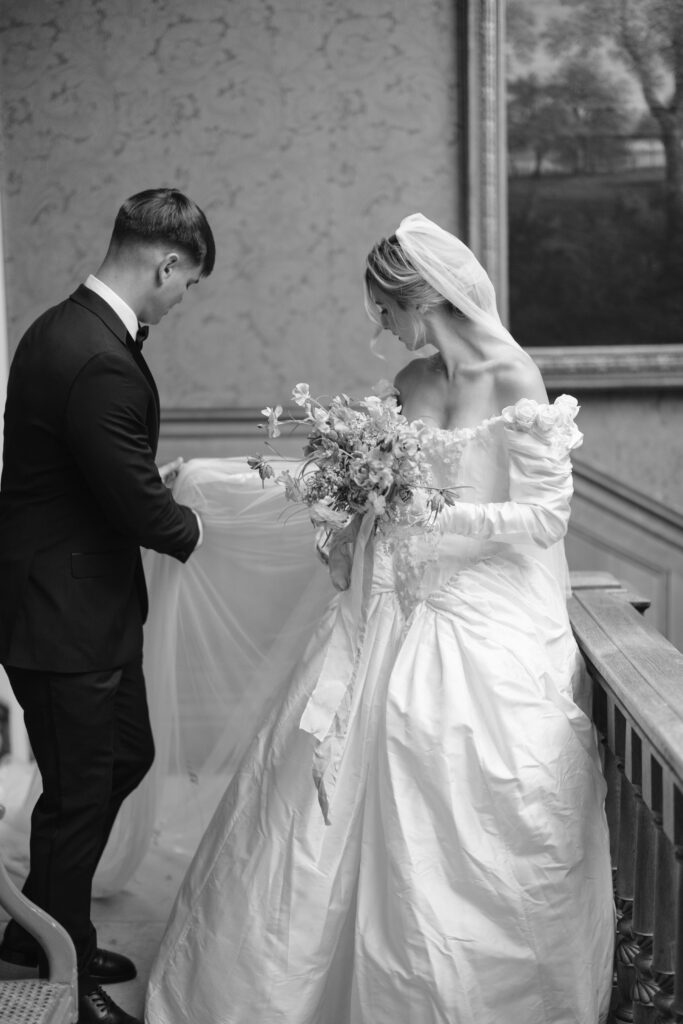 A bride in a white gown holding a bouquet stands beside a groom in a suit, who adjusts her veil. They are indoors by a wooden railing, with a painting on the wall behind them. Black and white photo.