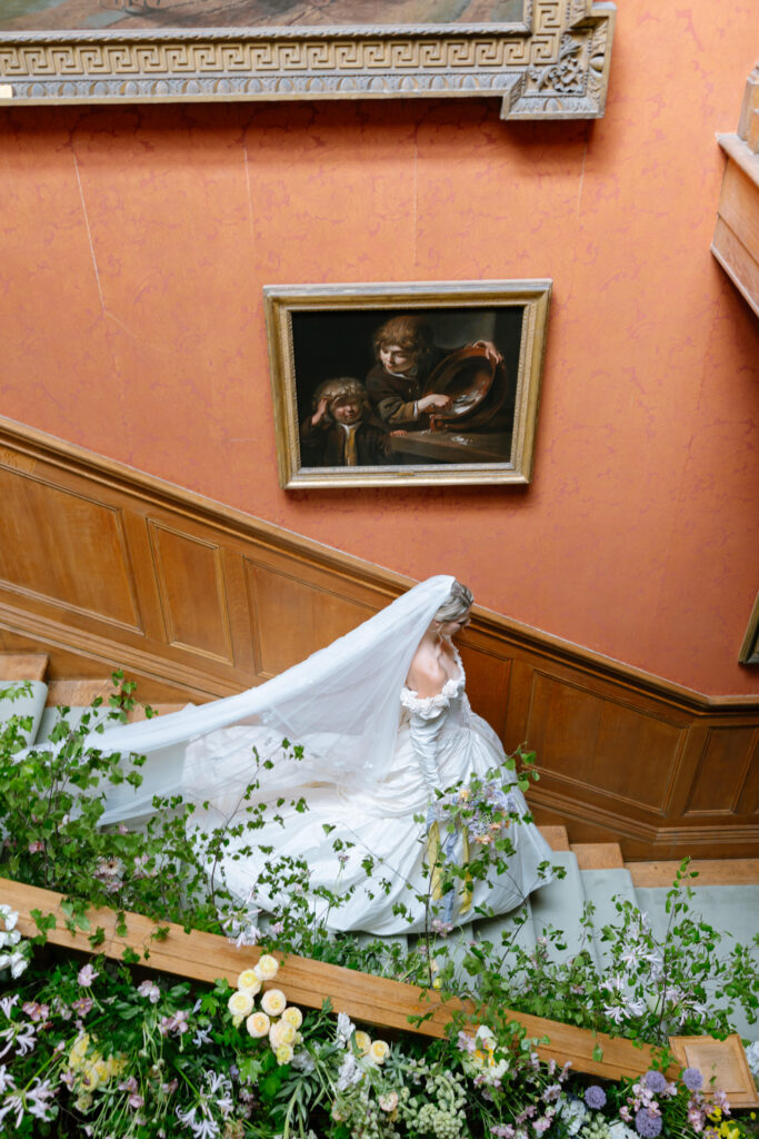 A bride in a white gown and long veil walks up a grand staircase decorated with flowers, beside a pink wall with a framed painting.