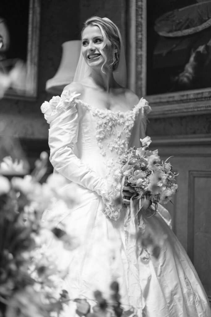 A smiling bride in a vintage-style wedding dress holds a bouquet. The black-and-white photo features soft lighting and floral details, with blurred flowers in the foreground and classical paintings in the background.