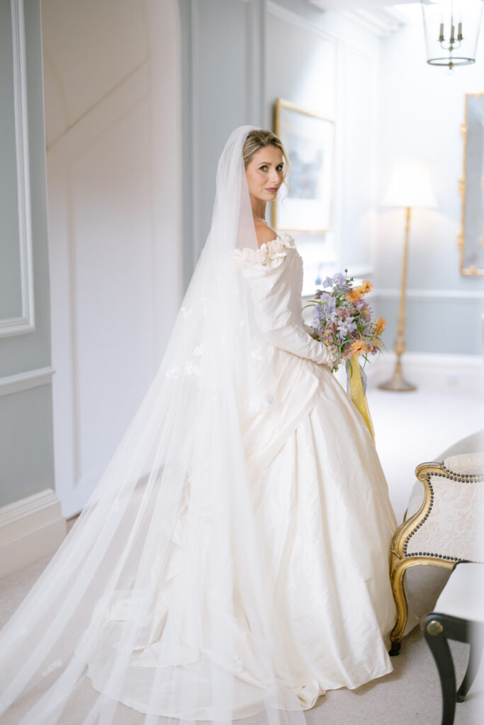 A bride in an elegant white gown and long veil holds a colorful bouquet while standing in a softly lit, classic room with light blue walls and ornate furnishings.