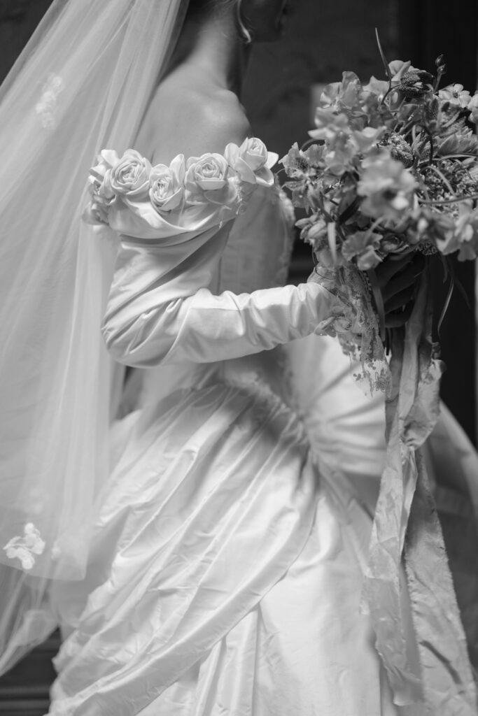 A bride in an off-the-shoulder wedding gown with floral details holds a bouquet of flowers. The image is in black and white.