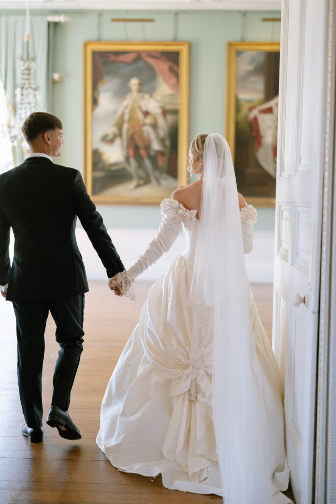 A bride in a white gown and veil holds hands with a groom in a black suit as they walk indoors, with large paintings and soft light in the background.