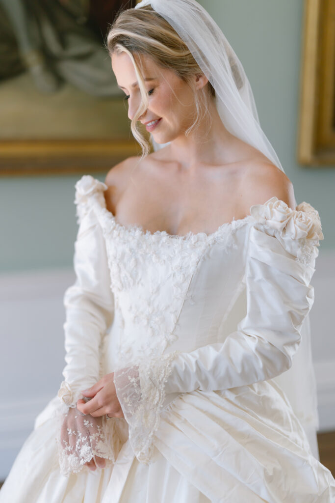 A smiling bride in an off-the-shoulder white wedding dress and veil stands indoors, looking down with her hands gently clasped.