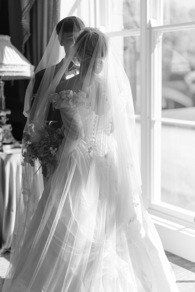 A bride and groom stand close together by a sunlit window, both wearing veils and formal wedding attire. The scene is captured in black and white, creating a soft and romantic atmosphere.