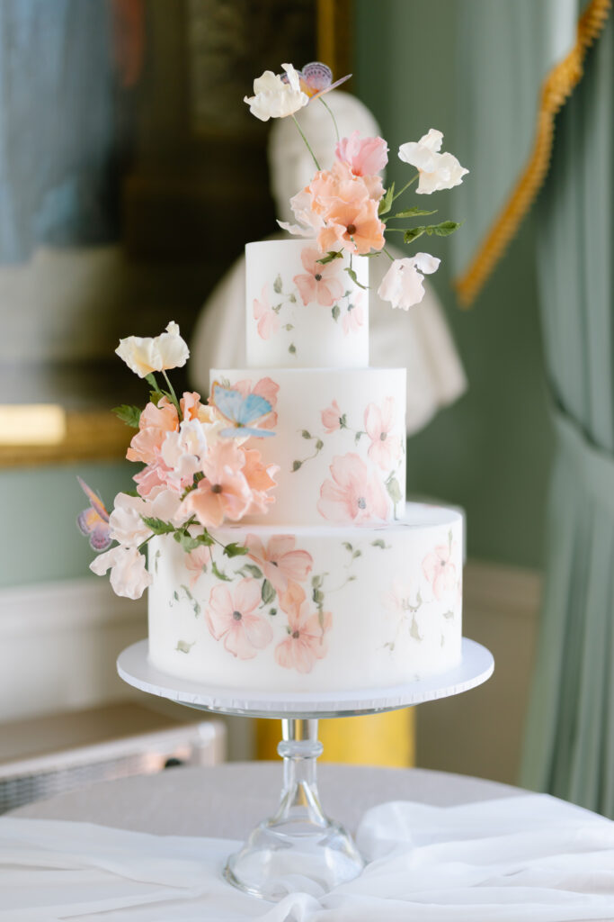 Three-tiered white cake decorated with pink and peach floral designs and topped with delicate, realistic sugar flowers, displayed on a glass cake stand in an elegant setting.