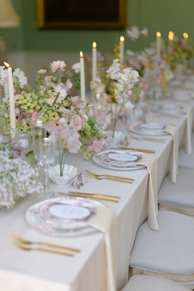 A long, elegant table set for a formal event features floral centerpieces, lit candles, gold flatware, glassware, and plates with menus, all arranged on a white tablecloth with cream napkins.