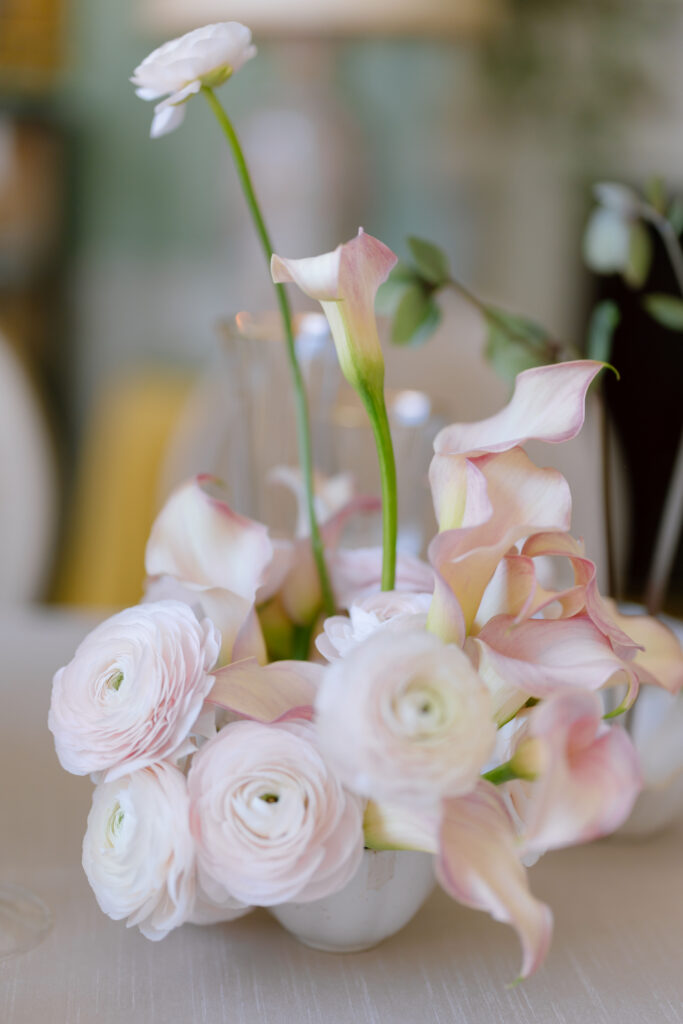 A white vase holds pale pink calla lilies and white ranunculus flowers, arranged delicately on a light-colored tablecloth.